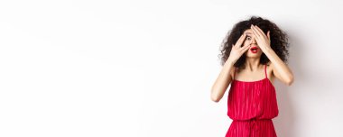 Heyecanlı kıvırcık saçlı, elbiseli, elleri ve parmaklarının arasından bakan, kameraya şaşırmış, beyaz arka planda duran.