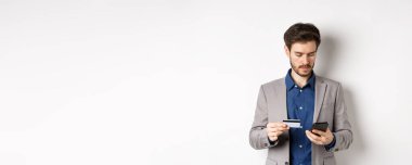 Çevrimiçi kredi kartı ve akıllı telefonla ödeme yapan yakışıklı bir işadamı, takım elbise içinde beyaz arka planda duruyor..