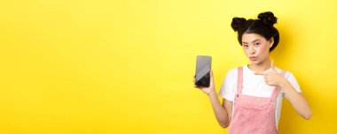 İnternetten alışveriş konsepti. Pembe makyajlı şık Asyalı kadın, boş akıllı telefonları işaret ediyor, iyi bir anlaşma gösteriyor, sarı arka plan..