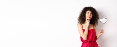 Kırmızı elbiseli düşünceli bir kız, küçük bir bulut tutuyor ve mutlu bir gülümsemeyle yukarı bakıyor, bir şeyler hayal ediyor, beyaz arka planda duruyor..