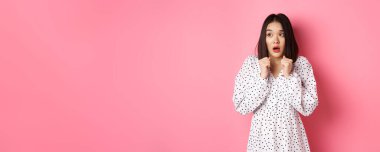 Korkmuş ve ürkek Asyalı kız korkudan titriyor, sola bakıyor ve soluk soluğa soluk soluğa, pembe arka planda dikilmiş duruyor..