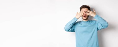 Şaşırtıcı gülümseyen adam elleriyle gözlerini kapatıyor ama beyaz arka planda duran muhteşem bir şeye parmaklarının arasından bakıyor..