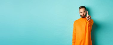 Ciddi sakallı adam kaşlarını çatıyor, hareket yasağı koymak için parmağını sallıyor, bir şeyi yasaklıyor, hayal kırıklığını dile getiriyor, açık mavi arka planda seninle aynı fikirde değil..