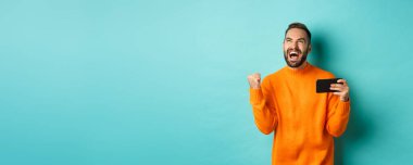 Cep telefonuyla kazanan adamın görüntüsü, yumruk tokuşturma ve zaferin sevinci, akıllı telefonu tatmin olmuş bir yüzle tutmak, turkuaz arka plan.