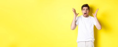 Hüsrana uğramış ve şok olmuş bir adam paniklemiş, bağırıyor ve korkmuş görünüyor. Sarı arka planda beyaz tişörtle dikiliyor..