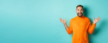 Heyecanlı adam reklam teklifine tepki veriyor, ellerini yana açıyor ve seviniyor, turkuaz arka planda turkuaz süveter giyiyor..