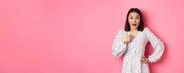 Güzel esmer Asyalı kadın kamerayı işaret ediyor. Seçilmiş kişi olarak şaşırmış görünüyor. Pembe arka plana karşı giyinmiş..