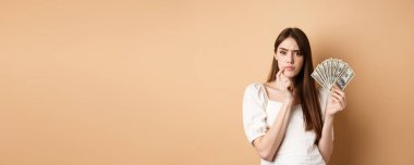 Ciddi ciddi para tutan ve düşünen, kameraya düşünceli görünen, kaşlarını çatan, dolar banknotlarıyla ne alacağına karar veren, bej arka plan.