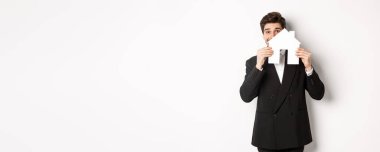 Siyah takım elbiseli yakışıklı simsar imajı, ev maketini gösteriyor ve gülümsüyor, ev satıyor, beyaz arka planda duruyor..