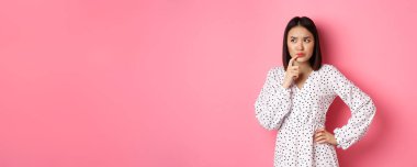 Sorunlu Asyalı kız karar veriyor, düşünürken kaşlarını çatıyor ve dudaklarına dokunuyor, sol üst köşede kararsız görünüyor ve seçiyor, pembe arka planda duruyor..