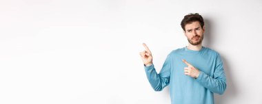 Şüpheci ve etkilenmemiş genç adam, yüzünü ekşitiyor, kötü terfiye bakıyor, beyaz arka planda tatminsiz duruyor..