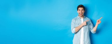 Heyecanlı, sıradan giyinmiş, reklamını yapan, fotokopi alanını işaret eden ve gülümseyen, mavi arka planda duran yakışıklı bir adam..