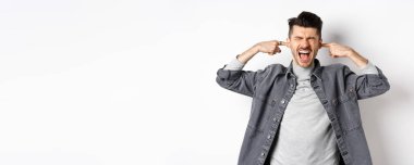 Sinirli adam kulaklarını kapattı ve çığlık attı, yüksek sesten rahatsız oldu, gürültülü müziği durdurmak için bağırdı, beyaz arka planda durdu..