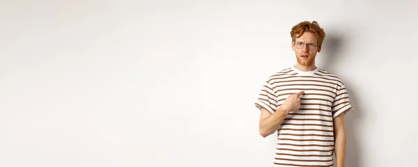Confused redhead man in glasses pointing at himself, looking questioned at camera, white background.