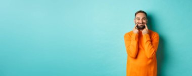 Sakallı bir adamın mutlu bir gülümsemeyle dudaklarını germesi, sahte mutluluk, açık mavi arka planda durması. Boşluğu kopyala