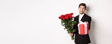 Tatil, ilişki ve kutlama kavramı. Siyah takım elbiseli, elinde bir buket kırmızı gülle sana hediye veren, beyaz arka planda duran yakışıklı bir adam..