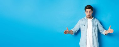 Heyecanlı yakışıklı adam büyük bir nesne gösteriyor ve şaşırmış görünüyor, mavi arka planda duruyor..