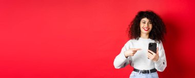 Akıllı telefonlu neşeli bir bayan. Telefonunu işaret ediyor. Tatmin olmuş bir şekilde gülümsüyor..