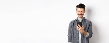 Yakışıklı adam telefonda mesaj okuyor ve gülümsüyor. Akıllı telefon tutan ve beyaz arka planda duran, ekrana bakan adam..