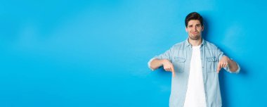 Hayal kırıklığına uğramış, sıradan kıyafetli, parmaklarını şüpheli bir suratla gösteren, mavi arka planda duran adam..