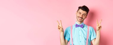 Papyon takmış, pantolon askısı takmış, kameraya gülümseyen, randevu için giyinmiş pembe arka planda duran romantik bir adam..