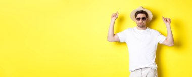 Turizm, seyahat ve tatil konsepti. Erkek turist tatilde, hasır şapka ve güneş gözlükleriyle dans ediyor, sarı arka planda poz veriyor..