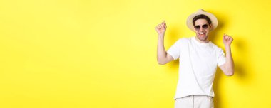 Turizm, seyahat ve tatil konsepti. Mutlu beyaz adam dans ediyor ve tatilde eğleniyor, güneş gözlüğü takıyor ve hasır şapka takıyor, sarı arka planda duruyor..