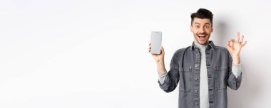 Heyecanlı gülümseyen adam boş akıllı telefon ekranı gösteriyor. İyi bir jest yapıyor, uygulama veya alışveriş öneriyor, beyaz arka planda duruyor..