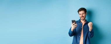 Şanslı adamın çevrimiçi kazandığı, cep telefonu ekranını okuduğu ve yumruk pompası yaptığı, zafer kazandığı, mavi arka planda durduğu görüntü.