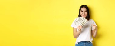 Genç Asyalı kadın gülümsüyor, ödül parasını gösteriyor, dolarları işaret ediyor, sarı arka planda duruyor..