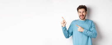 Mavi kazaklı mutlu bir beyaz adam, sol üst köşeyi işaret ediyor, beyaz arka planda bağlantı ya da logo gösteriyor, kameraya gülümsüyor..