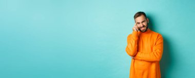 Sakallı, suratını buruşturan, utanç verici bir şeyden korkan, garip suratlı, açık mavi arka planda duran bir adam..