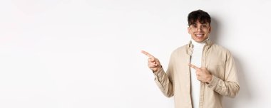 Gözlüklü neşeli yakışıklı adam gülümsüyor, parmaklarını logoya doğrultuyor, heyecanlı yüzüyle reklam yapıyor, beyaz arka planda duruyor..