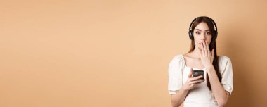 Şaşırmış genç bir kadın nefes nefese ve ağızını eliyle kapatıyor, kablosuz kulaklıkla podcast veya müzik dinliyor, elinde cep telefonu, bej arka plan.