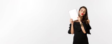 Moda ve alışveriş konsepti. Kırmızı dudaklı güzel bir kadın, siyah elbise giyiyor, dijital tablet gösteriyor ve öpüşme suratı yapıyor, beyaz arkaplan.