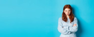 Kızgın kızıl saçlı, gücenmiş, göğsünde çapraz kollar ve somurtkan, kızgın bir şekilde kameraya bakan, mavi arka planda duran bir kız görüntüsü..