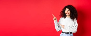 Heyecanlı, kırmızı dudaklı kıvırcık kadının portresi, logoyu şaşırmış bir şekilde gösteriyor, tanıtım anlaşmasına bakıyor, stüdyo arka planında duruyor..