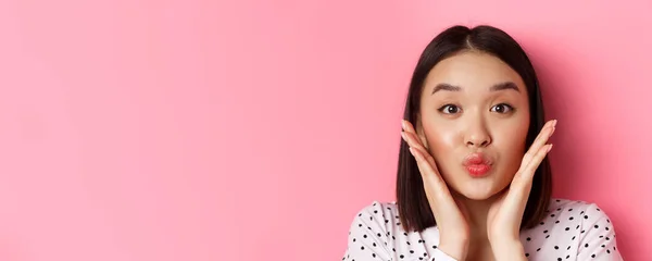 Beauty and lifestyle concept. Close-up of adorable asian woman touching face, pucker lips in kiss, standing over pink background.