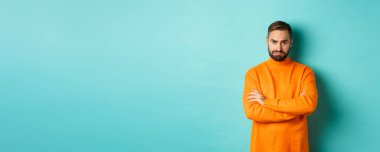 Aptal, üzgün, sakallı, somurtkan, gücenmiş, kolları çapraz göğsünde ve üzgün, açık mavi arka planda duran bir adam..