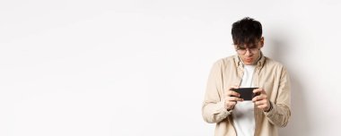 Yakışıklı genç adam mobil ekrana bakıyor ve akıllı telefondan video oyunu oynuyor, beyaz arka planda duruyor. Boşluğu kopyala