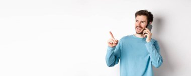 Teknoloji konsepti. Genç adam cep telefonuyla konuşuyor ve solu işaret ediyor, akıllı telefondan arıyor, beyaz arka planda duruyor..