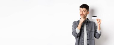 Düşünceli bir adam plastik kredi kartı tutuyor ve düşünüyor, kameraya bakıyor, seçim yaparken çenesine dokunuyor, beyaz arka planda duruyor..