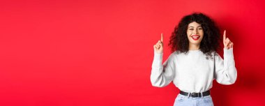 Kıvırcık saçlı mutlu genç bir kadın, parmaklarını havaya kaldırıp gülüyor, mükemmel beyaz bir gülümseme sergiliyor, kırmızı arka planda duruyor..
