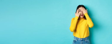 İlgi çekici Asyalı kız ilginç bir şeye parmak ucuyla bakıyor, kameraya bakıyor, mavi arka planda duruyor..