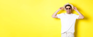 Mutlu adam tatilde, hasır şapka ve güneş gözlüğü takıyor, sarı arka planda dururken gülümsüyor..
