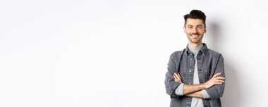 Kolları göğsünde çapraz gülümseyen yakışıklı genç adam, hazır hissediyor, kameraya bakıyor, beyaz arka planda duruyor..