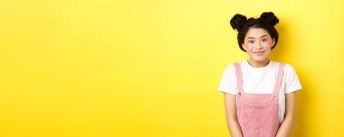 Makyajlı ve yaz elbiseli sevimli Asyalı kadın, kameraya aptal ve mutlu gülümsüyor, sarı arka plan..