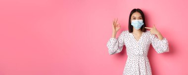 Coronavirus, sosyal uzaklık ve yaşam tarzı konsepti. Yüz maskesine bakan sevimli Asyalı kadın, tamam işareti gösteriyor, önleyici önlemler kullanarak destek veriyor, covid-19, pembe arka plan.