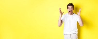 Öfkeli adam bağırıyor ve el sıkışıyor, nefretle yüzünü buruşturuyor, sarı arka plana karşı nefret dolu duruyor. Boşluğu kopyala