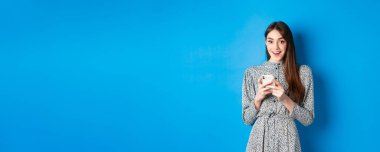 Uzun, doğal saçları olan, elbise giyen, akıllı telefon kullanan ve mutlu görünen, mavi arka planda duran tatlı bir kız..